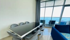 Un comedor moderno con una mesa rectangular y seis sillas, un sofá azul, y un ventanal con vistas al mar.