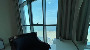 A room with a large window overlooking a beach, a black chair, and a glass table.