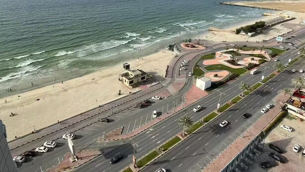 A coastal road with a beach on one side and a roundabout with palm trees.