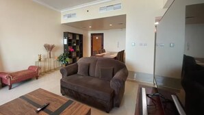 A spacious living room with a large brown sofa, a wooden coffee table, and a flat-screen TV. There is a bookshelf with various items and a dining area in the background.