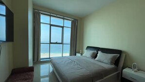 A bedroom with a large window offering a view of the beach, a bed with white bedding, a bedside table, and a bench.