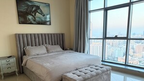 A bedroom with a large bed, a nightstand, a chair, a city view, and a painting on the wall.