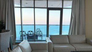 A modern living room with a large glass window overlooking the sea, a sofa, a glass coffee table, and a dining area with chairs.