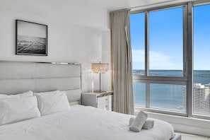 A hotel room with a large bed, a nightstand, a lamp, and a view of the ocean and buildings.