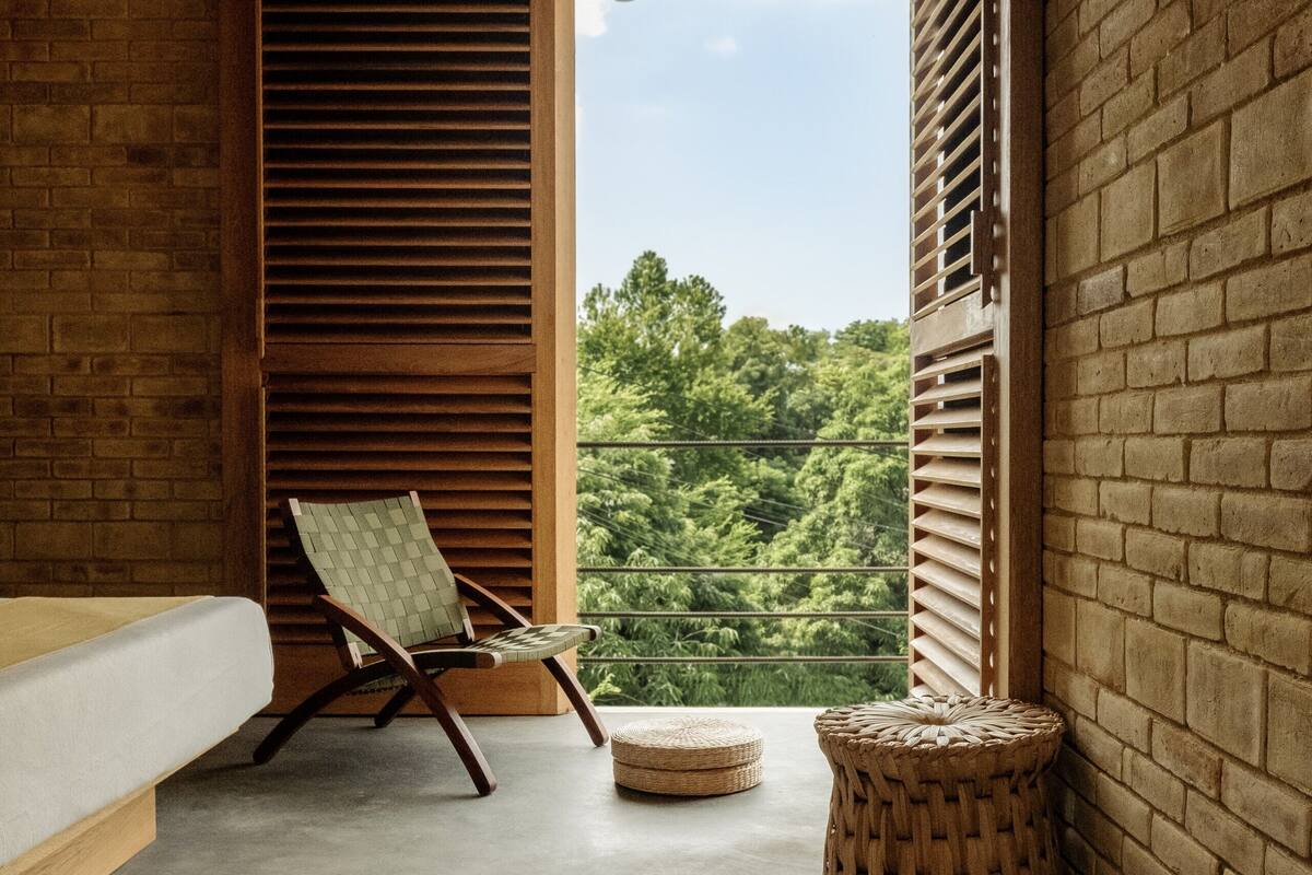 A balcony with a wooden chair, a woven stool, and a view of greenery.