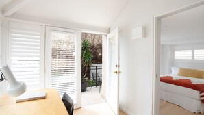 A bright, white interior with a wooden desk, a bed with a red and white blanket, and a balcony with a plant and a chair. - Petite Maison, Potts Point Isyd (Potts Point)