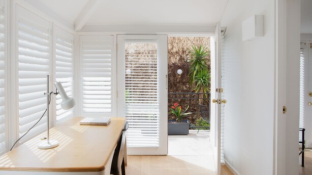 A bright room with white walls, a wooden table, and a desk lamp. There is a door leading to an outdoor area with plants and a fence. - Petite Maison, Potts Point Isyd (Potts Point)