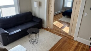 A living room with a dark sofa, a round coffee table, and a lamp. There is a wooden floor and a large mirror reflecting the room. - 5 Min to Salem~Comfy King Beds (Beverly)