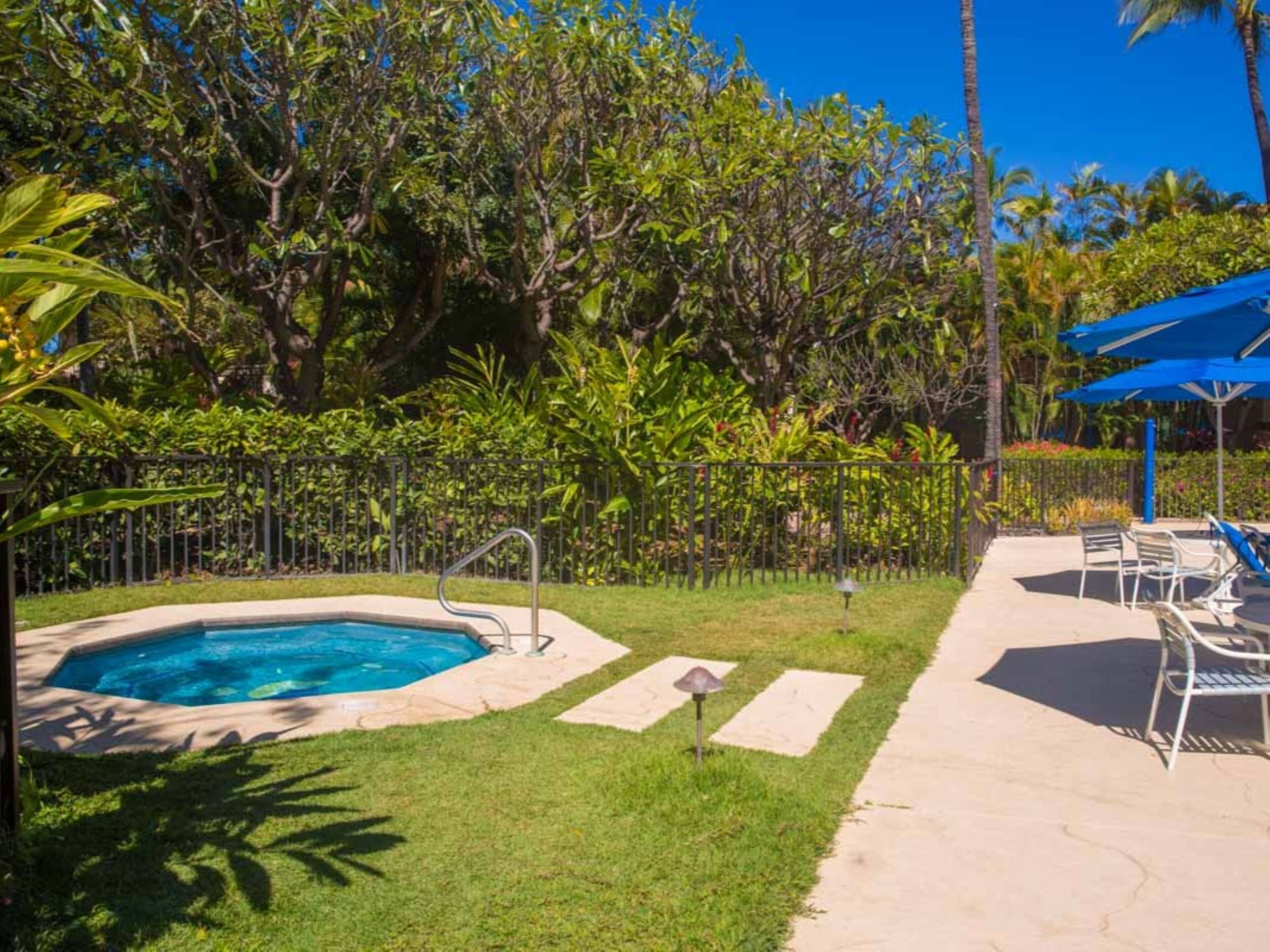 A swimming pool with a metal ladder, surrounded by a grassy area and a metal fence. There are lounge chairs under a blue umbrella.