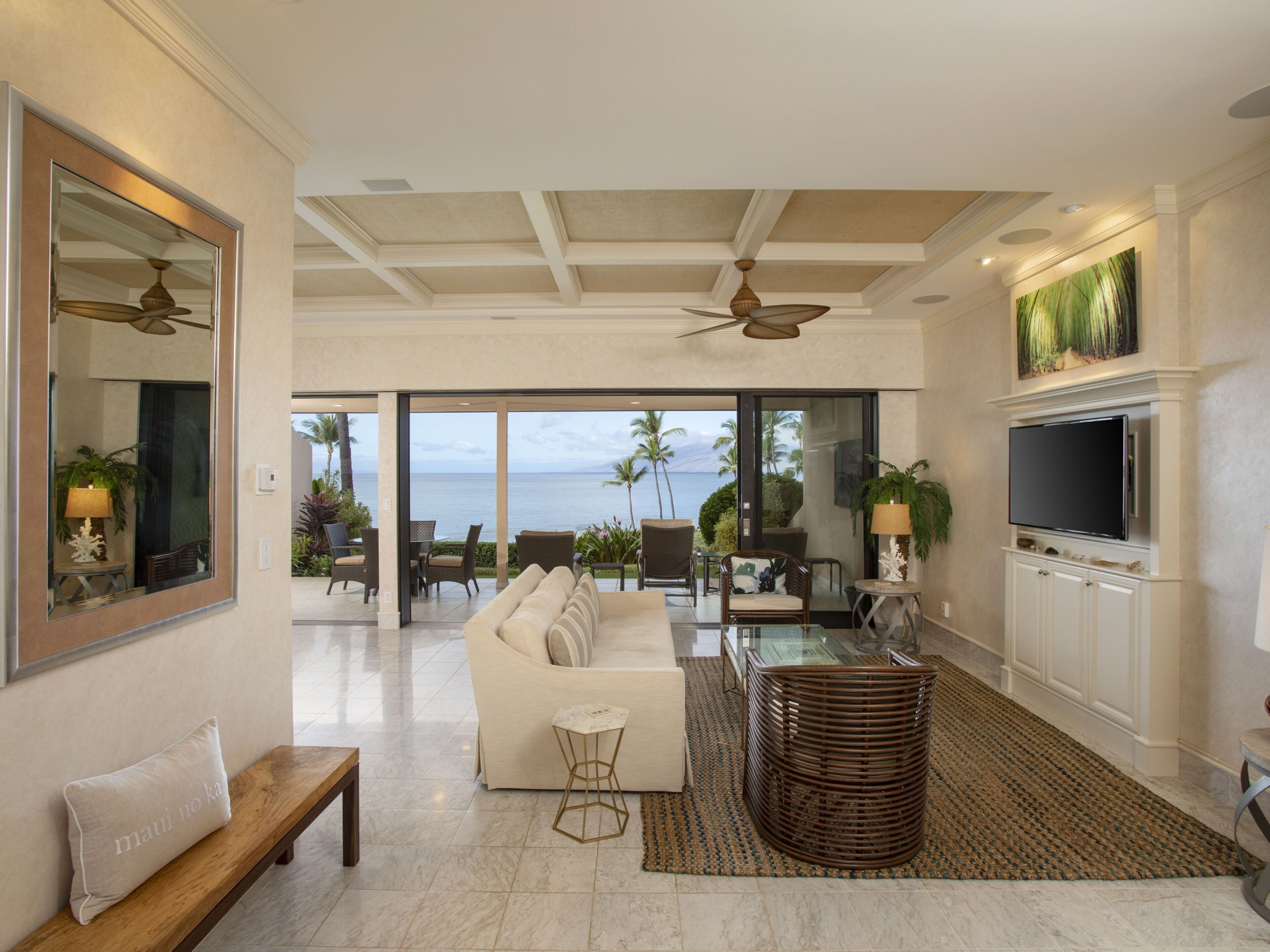 A spacious living room with a large sofa, a coffee table, and a TV mounted on the wall. There is a view of the ocean through large glass doors.
