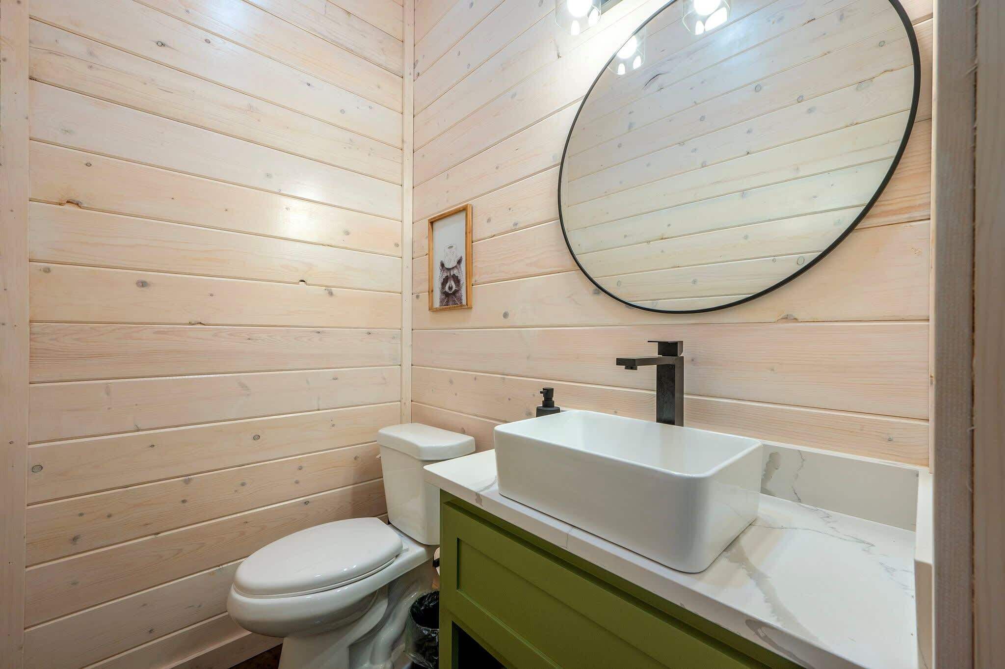 A bathroom with wooden walls, a white toilet, a white sink with a black faucet, and a round mirror.
