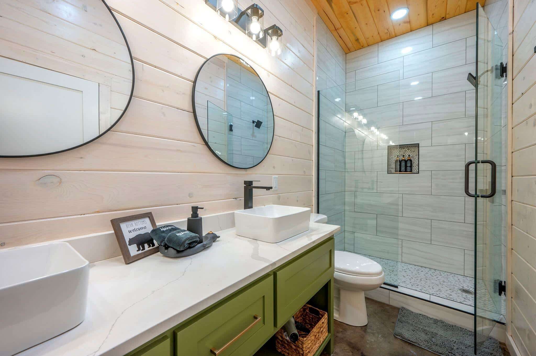 A modern bathroom with a wooden vanity, a large round mirror, a glass shower enclosure, and a toilet.