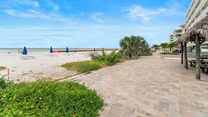 Ein Promenadweg am Strand mit Sonnenschirmen, einer Palme und einem Gebäude mit einem Reetdach.
