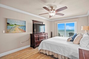 Une chambre avec un grand lit, un ventilateur de plafond, une commode, un tableau et une vue sur l’océan par une porte coulissante.