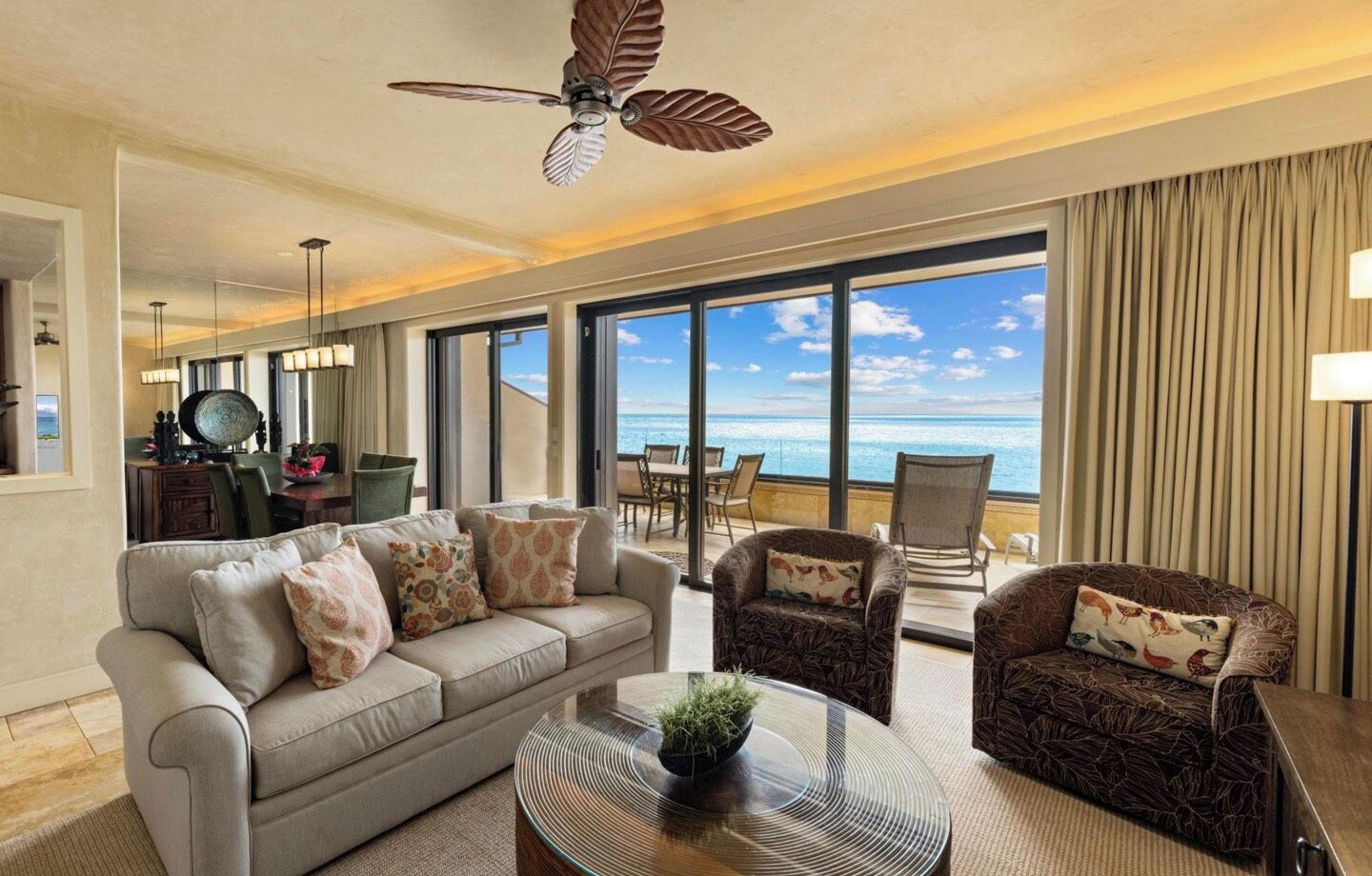 Une chambre d’hôtel dotée d’une grande fenêtre, d’un canapé, de deux fauteuils, d’un ventilateur de plafond et d’un coin repas offrant une vue sur l’océan.