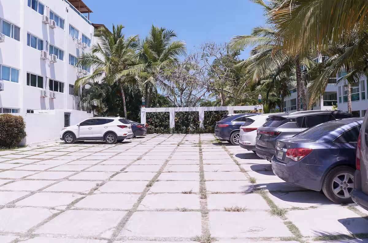 Un parking où sont garées plusieurs voitures, avec des palmiers et un bâtiment équipé de climatiseurs.