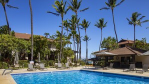 A swimming pool with lounge chairs and palm trees, surrounded by buildings with a tiled roof.