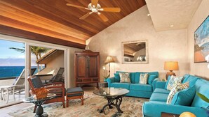 A spacious living area with a large sofa, a coffee table, and a dining table. There is a ceiling fan, a wooden cabinet, and a view of the ocean through large windows.