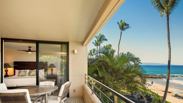 Ein Balkon mit einem Bett, einem Tisch und Stühlen mit Blick auf einen Strand, Palmen und das Meer.