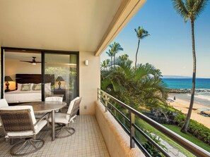 A balcony with a bed, a table, and chairs, overlooking a beach with palm trees and the ocean.