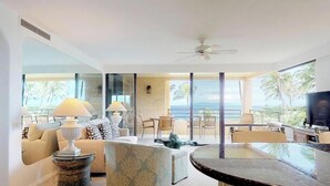 Una sala de estar moderna con un gran ventanal con vista al océano, un ventilador de techo y una zona de bar con televisor.