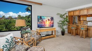A living room with a TV, wicker furniture, a ceiling fan, and a view of palm trees.
