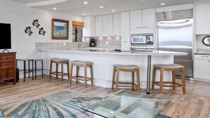 A modern kitchen with white cabinets, a stainless steel refrigerator, and a built-in oven. There are four wooden stools around a glass table.