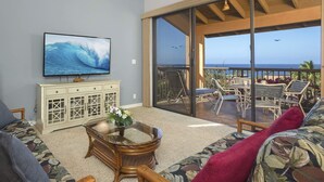Ein Wohnzimmer mit Fernseher, einem Glastisch und einer Couch. Es gibt einen Balkon mit Blick auf das Meer und Stühlen.