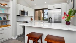 A modern kitchen with white cabinetry, stainless steel appliances, a large island with stools, and pendant lighting. - Platinum Rated and Professionally Managed (Wailea)