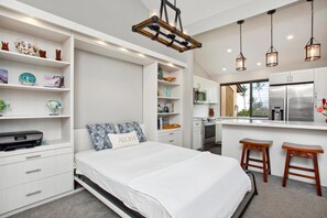 A compact bedroom with a bed, bedside tables, a desk, and a kitchenette.