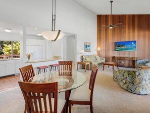 Una cocina moderna con armarios blancos, una zona de comedor con mesa de vidrio y sillas de madera, una sala de estar con sofá y televisor, y paneles de pared de madera.