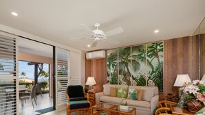 Una sala de estar con sofá, sillones, una mesa de centro y un comedor con mesa y sillas. Hay un ventilador de techo, un ventanal con persianas y un mural temático tropical.