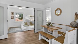 A room with a wooden floor, white walls, a white bench table with a bowl of fruit, and a white crib with a view of the outside. - Garden Home Estate, Two Kitchens (Portland)