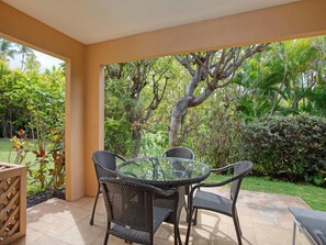 A covered patio with wicker furniture and a glass table, surrounded by lush greenery and a large tree. - Wailea Ekahi Village 3B | Garden-View Studio Near Pool & Beach (Wailea)