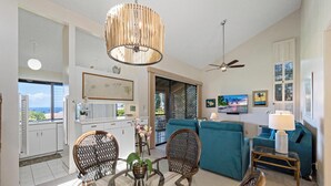A spacious living area with a dining table, chairs, and a large blue sofa. There is a ceiling fan, a TV mounted on the wall, and a view of the ocean through a window. - Wailea Ekahi Village 54D | Sweeping Ocean Views, Updated Kitchen & Resort Pools (Wailea)