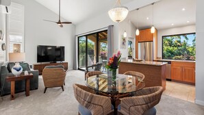 A modern living room with a glass dining table, wicker chairs, a flat-screen TV, and a ceiling fan. - Steps to Keawakapu Beach — Renovated Wailea Condo w/ Pool (Wailea)