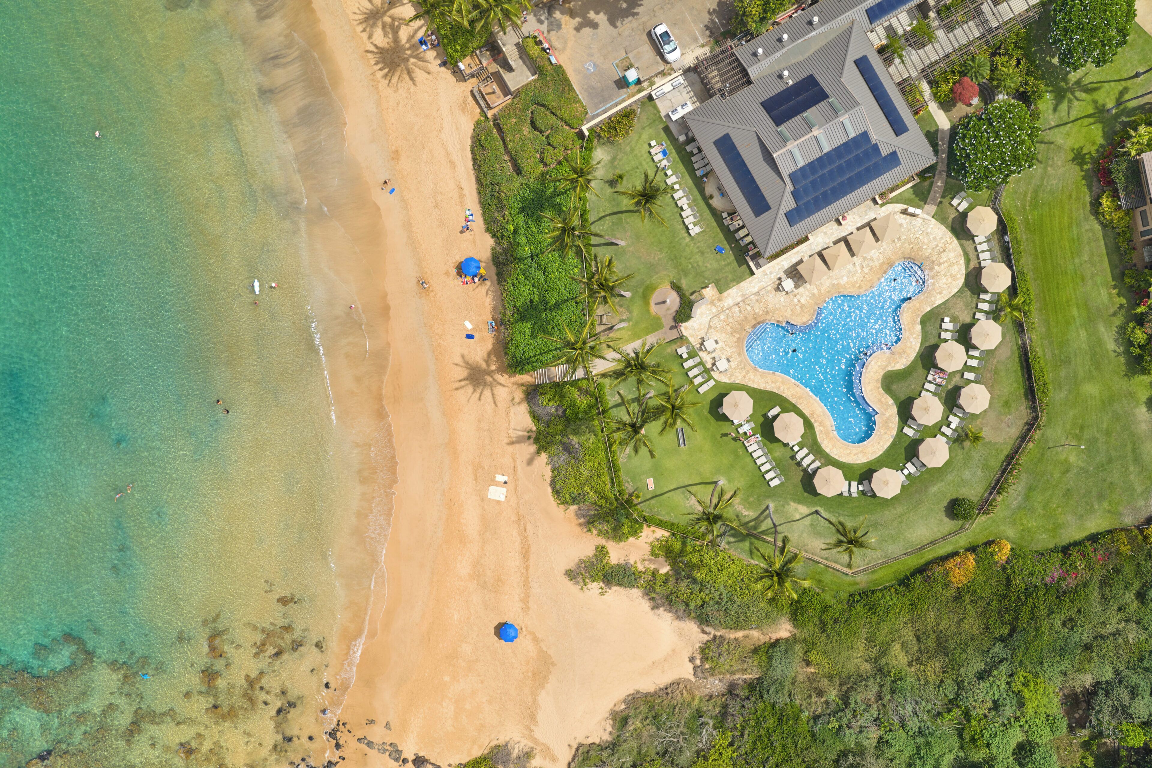 An aerial view of a resort with a beach, a swimming pool, and a building with a sloped roof.