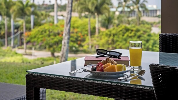 Une table avec une assiette de fruits, un verre de jus et une paire de lunettes de soleil, offrant une vue panoramique sur un paysage avec des palmiers et une étendue d’eau.