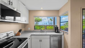 Una cocina moderna con armarios blancos, estufa de acero inoxidable, lavavajillas y ventana con vista a palmeras.