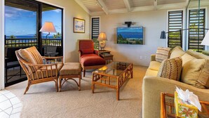 A spacious living room with a large window overlooking the ocean, a comfortable seating area with wicker chairs, a red armchair, and a sofa with patterned cushions.