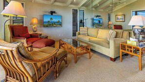 A spacious living room with a high ceiling, a large window, and a view of the ocean. There are two sofas, a coffee table, and a wicker chair.
