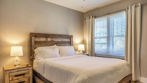 Ein Schlafzimmer mit Holzkopfteil, zwei Nachttischen mit Lampen, einem großen Bett mit weißer Bettwäsche und einem Fenster mit Jalousien.