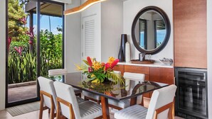 Un'area pranzo moderna con un tavolo in vetro, sedie di legno e vista su piante tropicali attraverso una porta a vetri.