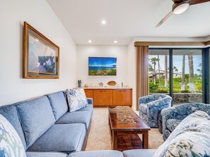A living room with a sofa, a coffee table, and patterned armchairs. There is a painting on the wall and a view of palm trees through the glass door.