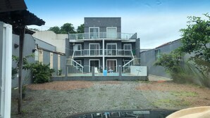 Un edificio de varios pisos con balcón, un patio de grava y vegetación circundante.