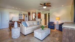 A spacious living room with a sofa, two ottomans, a coffee table, and a ceiling fan. - Wailea Elua Village 1301 | Front Row to the Ocean, Sunset Views & Steps to Ulua (Wailea)