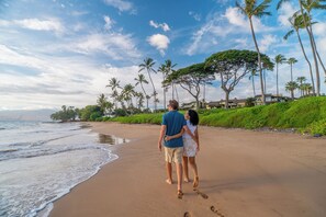Un couple se promène sur une plage de sable, bordée de palmiers, sous un ciel dégagé.