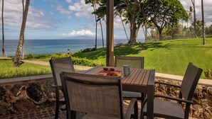 A covered outdoor seating area with a table and chairs, overlooking a golf course and the ocean. - Wailea Elua Village 1301 | Front Row to the Ocean, Sunset Views & Steps to Ulua (Wailea)
