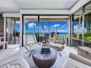 Een moderne woonkamer met een grote glazen deur die leidt naar een balkon met uitzicht op zee. Er staan twee witte fauteuils en een ronde salontafel met een plant.