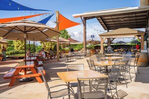 An outdoor patio area with tables and chairs, covered by umbrellas and a shaded structure.